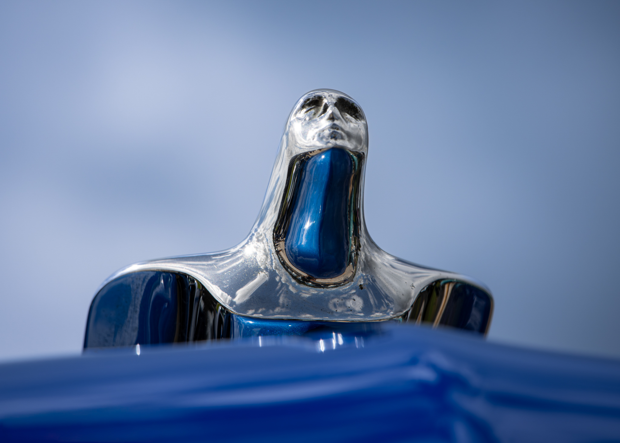 Blue and chrome vintage hood ornament photographed against a pale blue sky