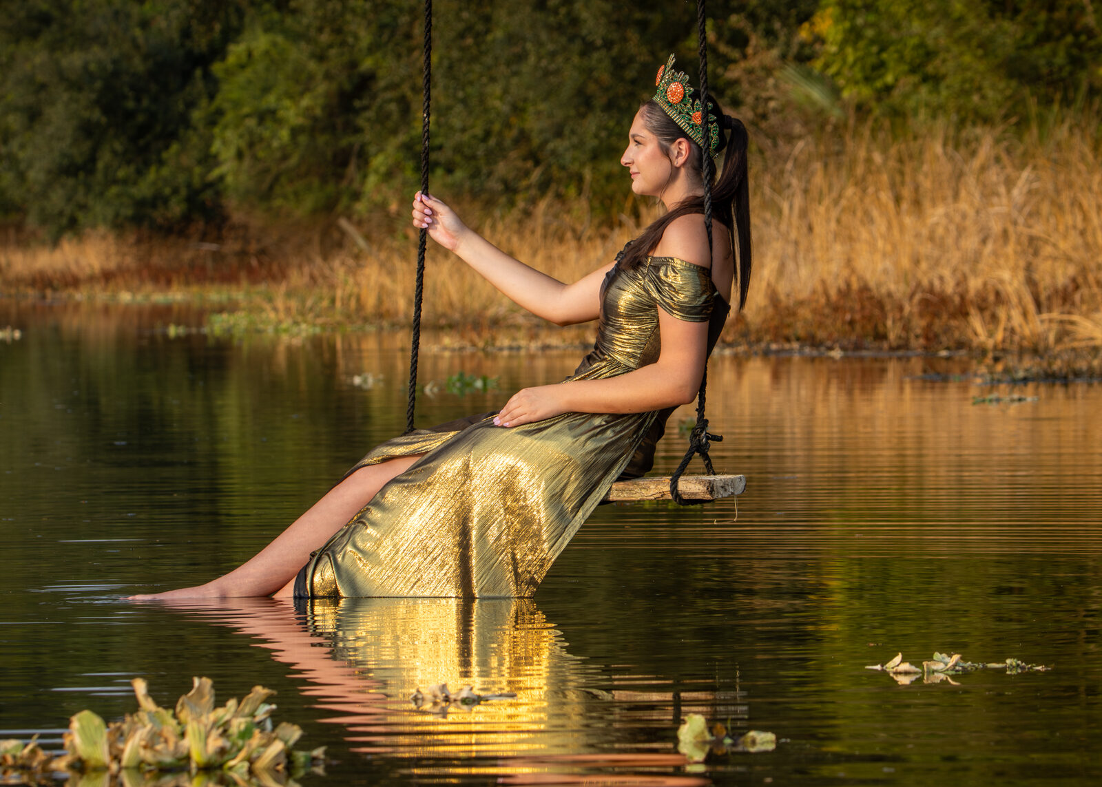 A crowned pageant portrait in a gold gown on a swing over water