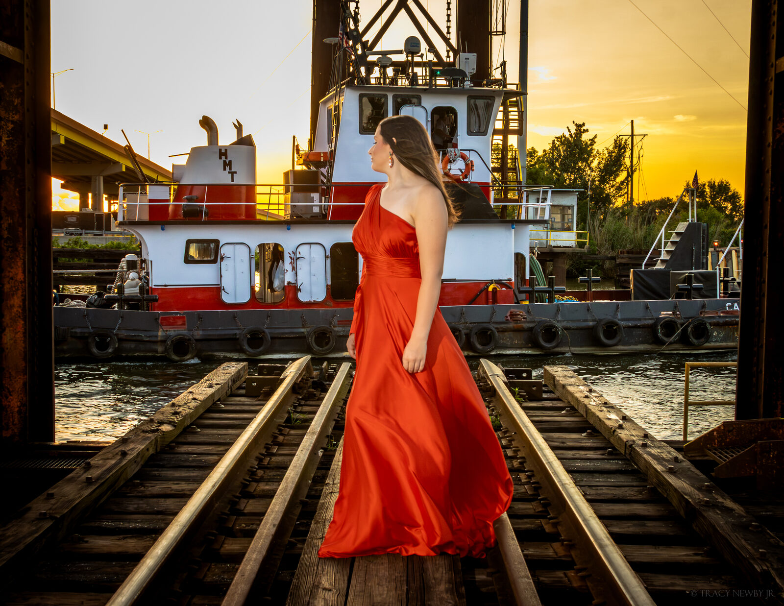 A pageant portrait in a red gown beside a working vessel at sunset
